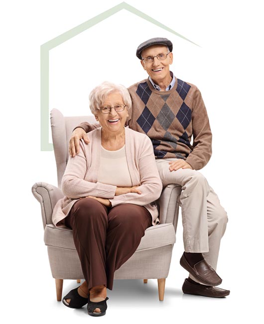 An elderly woman sits on an armchair with an elderly man standing beside her, both smiling. The man wears a cap and argyle sweater, while the woman wears glasses and a light pink cardigan.