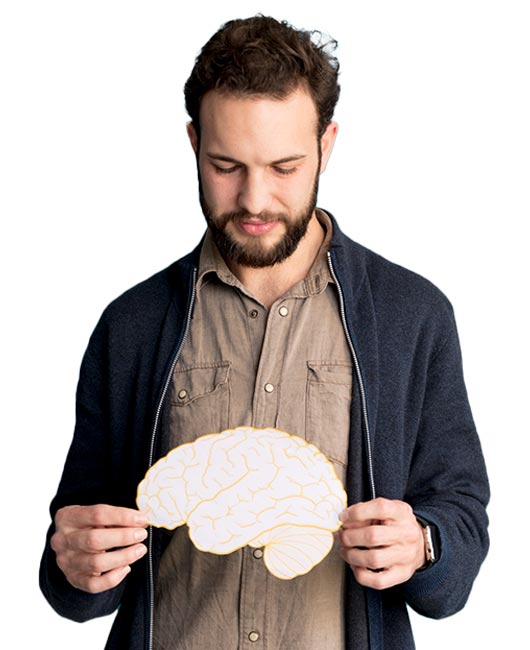 A man with a beard and casual clothing holds a paper cutout of a brain, looking down at it.