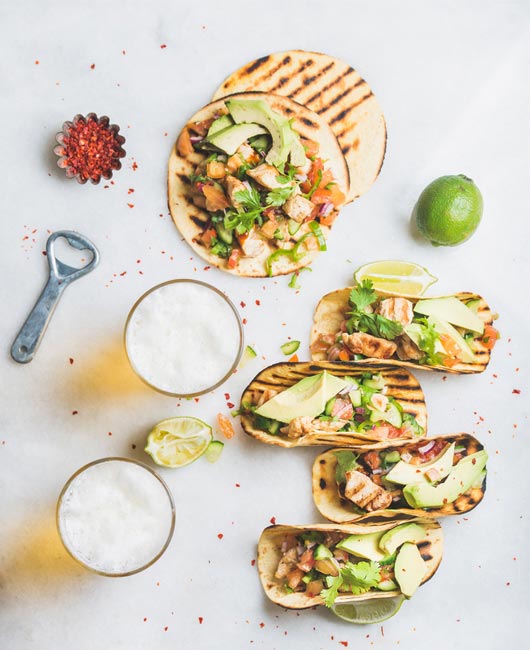 Grilled tortillas topped with diced vegetables, chicken, and avocado slices, accompanied by lime, a bottle opener, a slice of pomegranate, two glasses of beer, and scattered chili flakes.