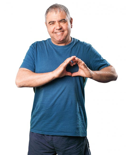 A man with short gray hair in a blue t-shirt and dark pants makes a heart shape with his hands while smiling.