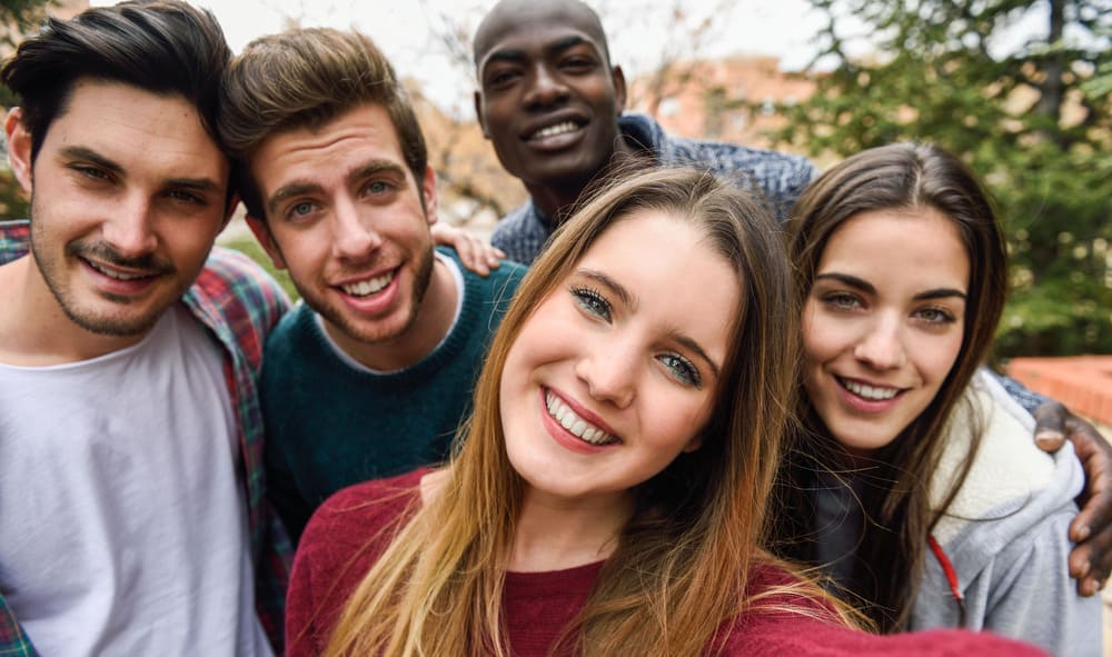 A group of five friends smiles at the camera for a selfie, outdoors in a natural setting with trees in the background.