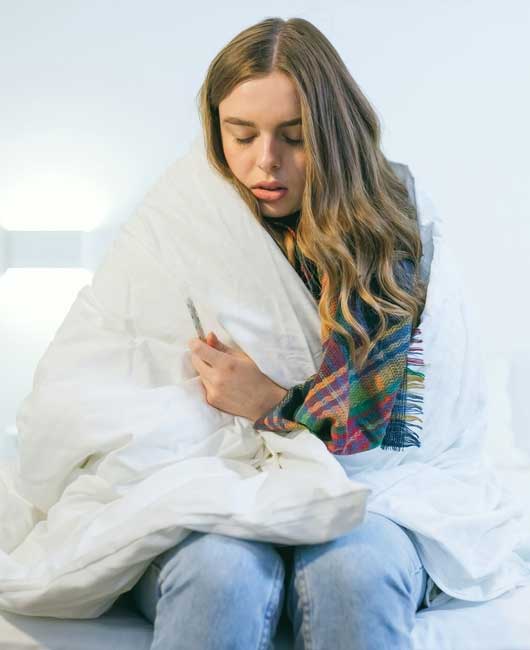 A woman with long hair sits wrapped in a white blanket, holding a thermometer and appearing unwell. She is wearing light blue jeans and a colorful scarf.