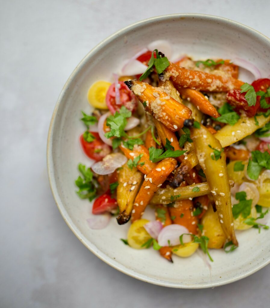 A bowl of roasted heirloom carrots garnished with sesame seeds and chopped parsley, accompanied by sliced radishes and cherry tomatoes.