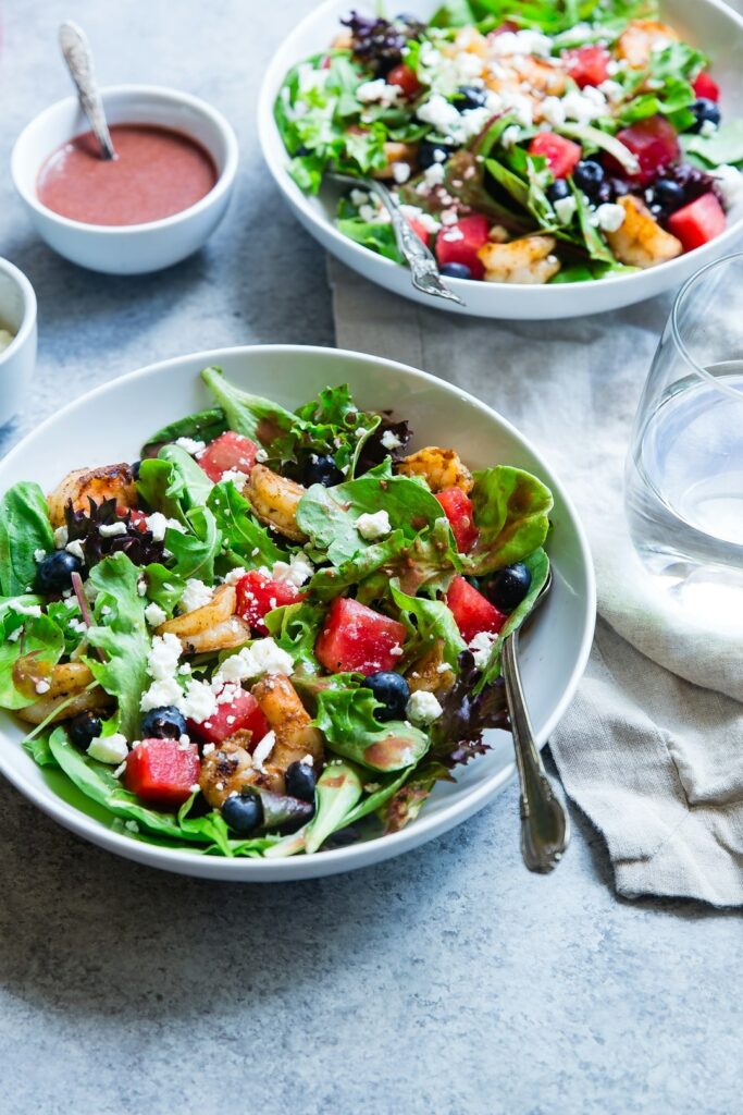 Two bowls of salad with mixed greens, watermelon, blueberries, grilled chicken, and feta cheese, with a fork in each bowl. Small bowls of dressing and a glass of water are nearby.