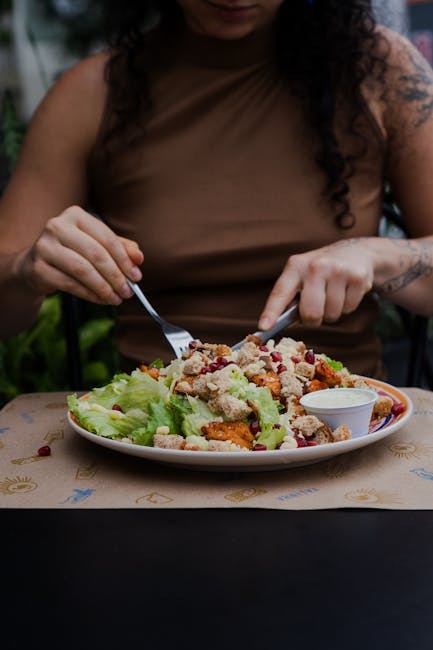 A person uses a fork and knife to eat a salad with lettuce, croutons, and a small cup of dressing on a plate.