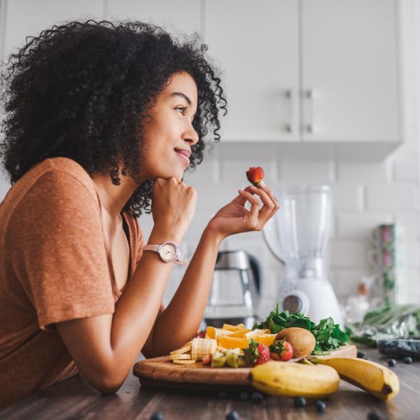 Woman sitting at a kitchen counter, holding a strawberry and looking away, with a cutting board of assorted fruits and a blender in the background.