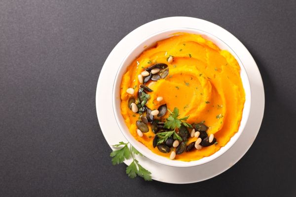 Bowl of mashed sweet potatoes topped with pumpkin seeds, pine nuts, herbs, and a sprig of parsley, on a dark surface.