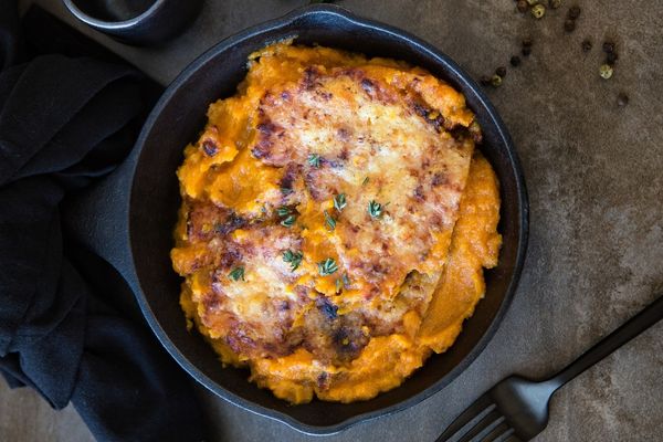 A cast iron skillet filled with baked cheesy mashed sweet potatoes, garnished with herbs, sits on a dark surface with a fork and black cloth nearby.