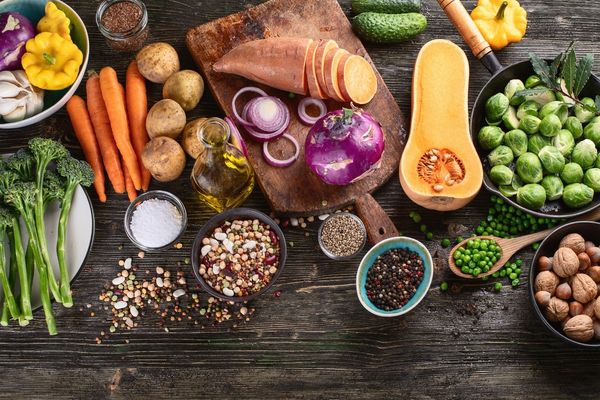 Assorted fresh vegetables, beans, nuts, spices, and oil arranged on a rustic wooden table, viewed from above. A cutting board holds sliced sweet potato, onion, and kohlrabi.