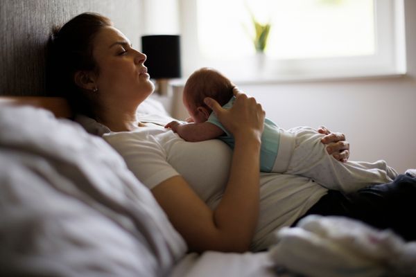 A woman lies in bed holding a baby on her chest. Both appear relaxed in a softly lit bedroom with a window in the background.