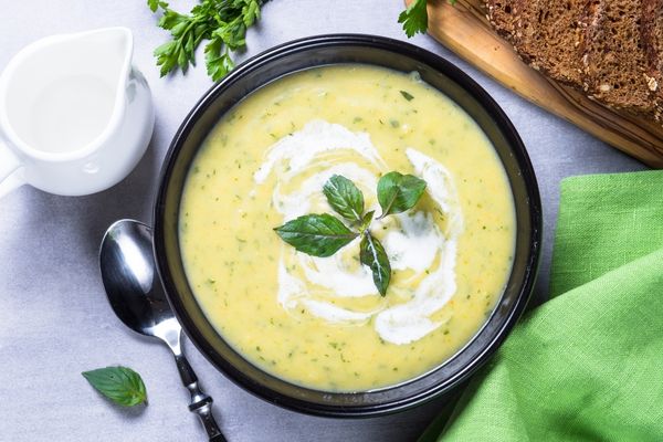 A bowl of creamy yellow soup with herbs, topped with a swirl of cream and basil leaves, next to a silver spoon, bread, and a green napkin.