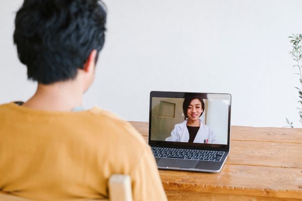 A person sits at a wooden table having a video call with a doctor on a laptop.