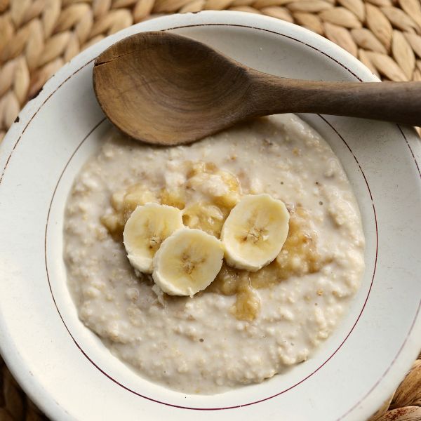 A bowl of oatmeal topped with banana slices, with a wooden spoon resting on the rim, sits on a woven placemat.