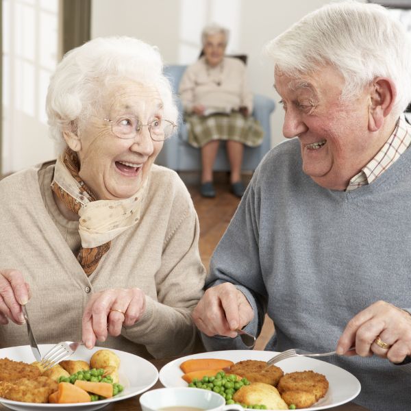 Elderly man and woman smiling and eating a meal together at a table, with another elderly woman seated in the background.