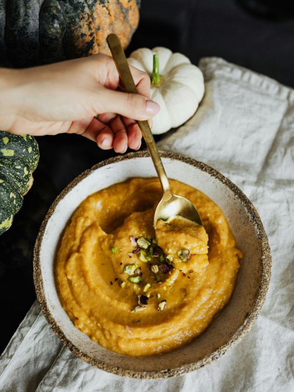 A hand holding a spoon scoops orange pumpkin soup from a bowl, garnished with chopped nuts. A white napkin and small pumpkins are in the background.