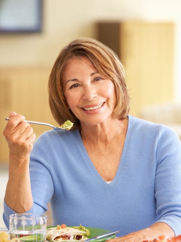 Older woman in a blue sweater smiles while holding up a forkful of food, seated at a table with a plate and glass in front of her.