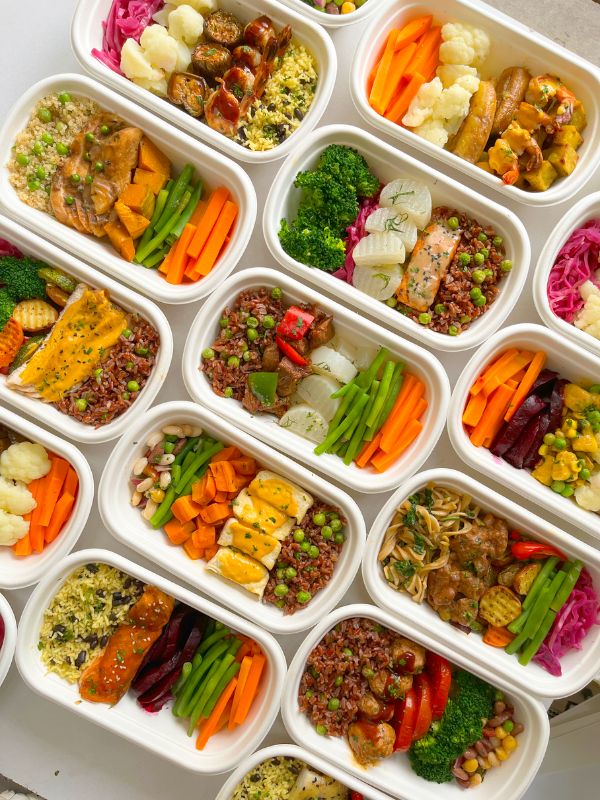 Several rectangular trays filled with assorted colorful vegetables, grains, and proteins arranged in a grid on a white surface.