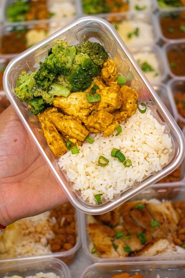A hand holds a meal prep container with white rice, seasoned chicken, and broccoli, garnished with chopped green onions. Other similar meal containers are visible in the background.